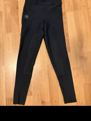 Svarta ridleggings från Alequi - Snygga svarta ridtights från Alequi med helskoning i silikon för extra grepp. Tightsen har hög midja och Alequi-logga på låret. Tillverkade i stretchigt funktionsmaterial som passar perfekt för ridning och träning.Jättefina Alequi ridbyxor i storlek xs med snygg passform.  Ridbyxorna var silikon på insidan av midjan som håller byxorna på plats  Bild 2/3 mobilficka. Små hål.  Bild 4 helskoning. Något skavt men fyller fortfarande sin funktion   Bild 7 sömmar vid båda ben slut. Inga hål bara sömnen