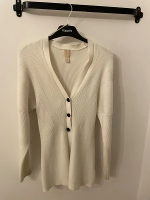 Vit ribbad kofta med knappar - Stilren vit kofta från HM med ribbad struktur och djup V-ringning. Koftan har tre svarta knappar framtill och långa ärmar. Materialet är mjukt och stretchigt, vilket ger en bekväm passform. Perfekt att slänga över en topp för en enkel men snygg look.