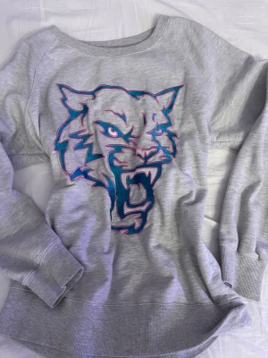 Grå sweatshirt med tigertryck - Cool grå sweatshirt från Westhill Redbird med ett stort tigertryck i blått och rosa på framsidan. Tröjan har rund halsringning, långa ärmar och ribbade muddar. Perfekt för en avslappnad och trendig stil.