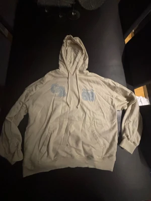 Beige hoodie med tryck CA 98 - Snygg beige hoodie med huva och känguruficka framtill. På bröstet står det 'CA 98' och 'Oceans City' i ljusblått tryck. Hoodien har dragsnören vid huvan och ribbade muddar vid ärmar och nederkant. Perfekt för en avslappnad och trendig look.