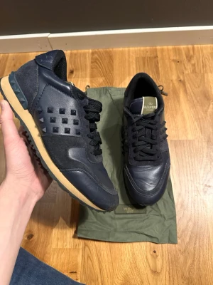 Valentino Rockrunner sneakers marinblå - Snygga marinblå Valentino Rockrunner sneakers med ikoniska nitar på sidan. Skorna har ovandel i skinn och mocka, svarta snören och beige sula. Perfekta för dig som vill ha en unik och trendig look.