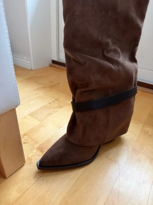Bruna mockastövlar med spetsig tå - Säljer ett par bruna stövlar i mocka med spetsig tå och låg klack. Stövlarna har en slouchy passform och ett svart band runt vristen som detalj. Perfekta för dig som vill ha en trendig och unik look.