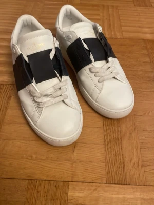 Valentino Garavani vita sneakers - Snygga vita sneakers från Valentino Garavani med bred marinblå detalj över snörningen och klassiska nitar på hälen. Skorna har rund tå, platt sula och är tillverkade i skinn. Perfekta för dig som vill ha en stilren och exklusiv look.