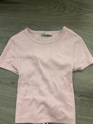 Ljuslila ribbad topp med snörning - Säljer en ljuslila ribbad topp från PULL&BEAR i storlek S. Toppen har korta ärmar och en snygg snörning i ryggen som ger en unik look. Materialet är mjukt och stretchigt, perfekt för dig som gillar trendiga detaljer.