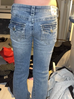 True Religion blå bootcut jeans - Säljer ett par blå jeans från True Religion med bootcut passform. Jeansen har dekorativa bakfickor med lock, knappar och blingdetaljer. Klassisk femficksmodell, normal midja och snygg tvättad look. Modellen är joey asså väldigt lågmidjade använda max 3 gånger helt nya säljer då pågrund dom inte kommer till användning ni får jätte Järna lägga bud! Och fråga om ni vill veta mer.