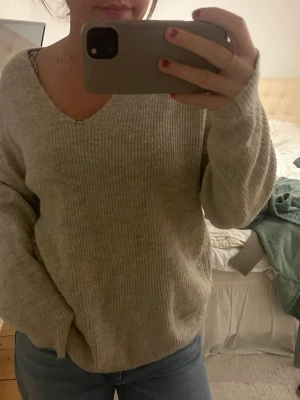 Beige ribbstickad tröja med v-ringning - Mysig beige stickad tröja med v-ringad hals och avslappnad passform. Tröjan har ribbstickad struktur och långa ärmar, perfekt att matcha med jeans för en chill look. Inget synligt märke eller logga.