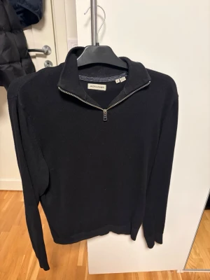 Svart halvzip-tröja Jack & Jones - Svart stickad tröja från Jack & Jones med halvzip och hög krage. Tröjan har lång ärm och ribbade muddar vid ärmslut och nederkant. Enkel och stilren design som passar till det mesta. Perfekt för lager på lager under kyliga dagar. Storlek M, inga skador eller sådant!