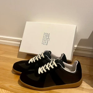 Svarta Maison Margiela sneakers - Maison Margiela sneakers i svart med kontrasterande vita snören och gummisula. En rund tå och platt sula. Klassisk sportig siluett med diskret logotyp på tungan och vit insida. Perfekt för dig som gillar stilrena och lyxiga sneakers. Box ingår.