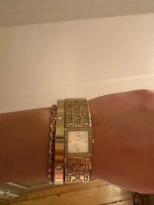 Guldig vintage klocka från Guess - Säljer en snygg guldig klocka från Guess med fyrkantig urtavla och detaljerade länkar. Klockan har glittriga stenar runt urtavlan och ett coolt mönster på armbandet. Perfekt accessoar för att lyfta din outfit.