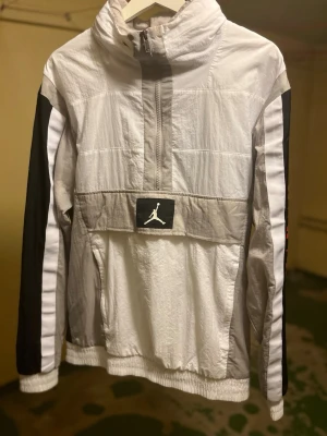 Jordan vindjacka i vitt och svart - Jordan vindjacka i vitt och svart med röda detaljer på ena ärmen. Jackan har halv dragkedja framtill, hög krage och elastiska muddar. Används inte längre därför säljas.