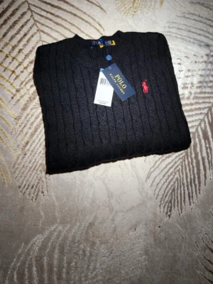 Svart kabelstickad tröja Polo Ralph Lauren - Kabelstickad svart tröja från Polo Ralph Lauren med klassisk röd logga broderad på bröstet. Tröjan har rund halsringning och ribbade muddar. Perfekt för dig som gillar stilrena och tidlösa plagg.