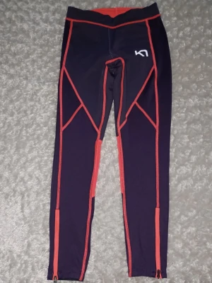Svarta och röda träningstights Kari Traa - Snygga träningstights från Kari Traa i lila med röda detaljer och sömmar. Tightsen har dragkedjor vid bensluten och logga på både fram- och baksidan. Tillverkade i stretchigt funktionsmaterial som passar perfekt till träning. Etiketter bortklippta då jag alltid tar bort dem när jag får hem nytt.  Nyskick.