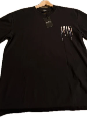 Svart AMIRI t-shirt  - Svart t-shirt från AMIRI med coolt tryck av märkesnamnet och färgstänk i vitt, blått och orange både fram och bak. Klassisk passform och rund halsringning. Perfekt för dig som vill sticka ut med en unik streetwear-look.