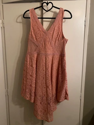 Rosa spetsklänning med v-ringning - Söt rosa klänning i spets med v-ringning både fram och bak. Klänningen är ärmlös och har fina spetsdetaljer samt transparent mesh i sidorna. Dragkedja i sidan för enkel påklädning. Perfekt för sommarens alla tillfällen!