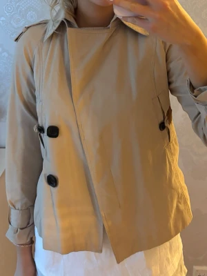 Kort beige trenchcoat med knappar - Snygg kort trenchcoat i beige med dubbelknäppning och axelklaffar. Jackan har långa ärmar med remmar vid ärmsluten och klassisk krage. Jag är 180cm lång och bär vanligtvis S/M. Skicka gärna frågor eller prisförslag!