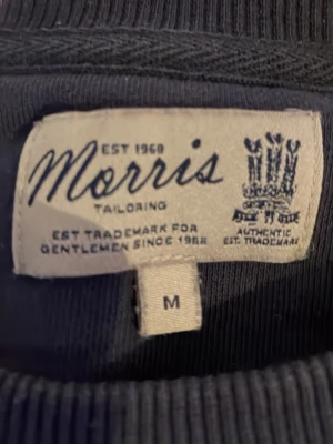 Blå sweatshirt från Morris - Mörkblå sweatshirt från Morris med tryck 'Morris Gentlemen's Club' i vitt på bröstet. Tröjan har rund halsringning, ribbade muddar vid ärmslut och nederkant samt en klassisk, avslappnad passform. Perfekt för en stilren och bekväm look.
