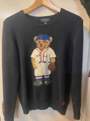 Mörkblå stickad tröja Polo Bear 14-16 - Mörkblå stickad tröja från Polo Ralph Lauren med den ikoniska Polo Bear i basebolloutfit på bröstet. Tröjan har rund halsringning, långa ärmar och ribbade muddar vid ärmslut och nederkant. Perfekt för dig som gillar klassiska och lekfulla detaljer.