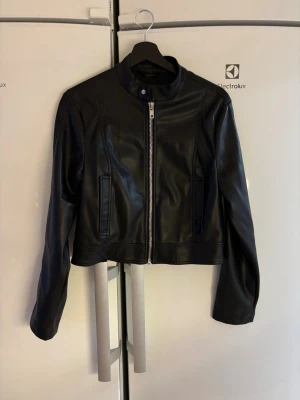 Svart croppad skinnjacka - Snygg svart skinnjacka med croppad passform och raka linjer. Jackan har en hög krage, silverfärgad dragkedja framtill och två fickor. Perfekt för dig som vill ha en stilren och edgy look.