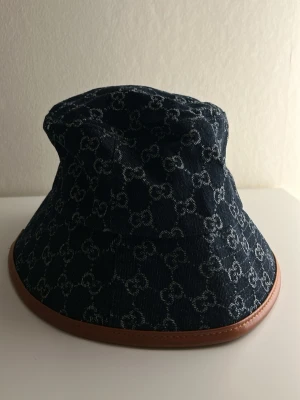 Bucket hat i denim - Snygg bucket hat i mörkblå denim med klassiskt GG-mönster och brun läderkant. Hatten har en avslappnad form och är fodrad på insidan för extra komfort. Perfekt accessoar för att lyfta din stil med en lyxig touch.