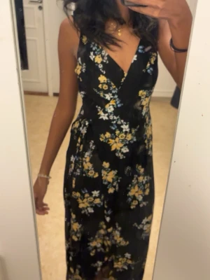 Svart blommig omlottklänning - Säljer en svart omlottklänning med blommigt mönster i gult och vitt. Klänningen har v-ringning och smala axelband, samt en figurnära passform som ger en snygg siluett. Perfekt för sommarens alla tillfällen!