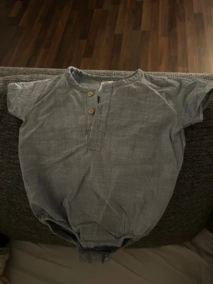 Bebis kläder Grå kortärmad body från H&M - Kortärmad body i grått från H&M, tillverkad i ekologisk bomull. Modellen har rund hals, tre träknappar framtill och tryckknappar i grenen för enkel påklädning. Tunn och mjuk kvalitet, perfekt för små barn.