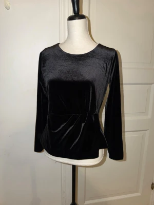 Svart sammetsblus med twistdetalj - Stockh LM - Säljer en svart långärmad blus i mjuk sammet. Toppen har rund halsringning och en snygg twistdetalj i midjan som ger en smickrande siluett. Perfekt för dig som gillar stilrena plagg med lite extra känsla. Ingen synlig logga eller märke.