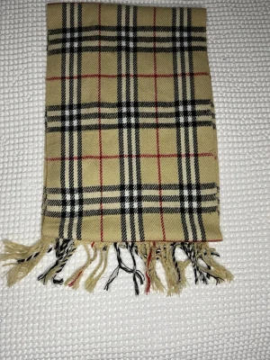 Beige rutig halsduk med fransar burberry - Klassisk beige halsduk med rutigt mönster i svart, vitt och rött. Halsduken har fransar i ändarna och är tillverkad i ett mjukt tyg som känns skönt mot huden. Perfekt accessoar för att lyfta din outfit med en tidlös look.