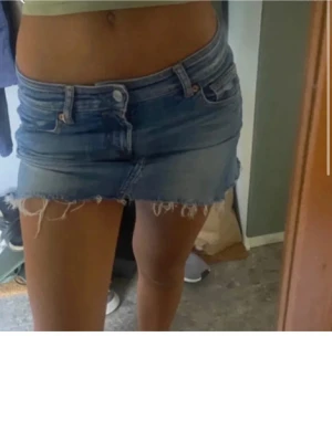 Blå jeanskjol med fransig kant - Säljer en kort blå jeanskjol med rå, fransig kant och klassiska fickor fram och bak. Kjolen har en låg midja och stängs med knapp och dragkedja. Perfekt för dig som gillar en avslappnad och trendig stil.