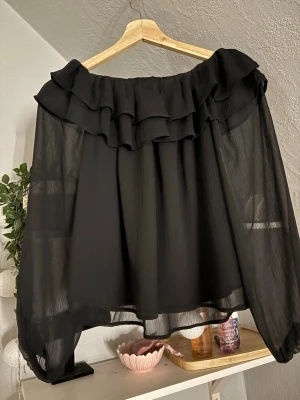 Svart offshoulder blus med volang - Trendig svart offshoulder blus i transparent, luftig chiffong med bred volang runt axlarna. Perfekt för dig som gillar en romantisk och bohemisk stil. Blusen har vida, långa ärmar och en lös passform som ger en avslappnad vibe.