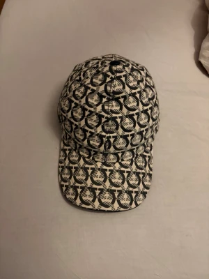 Keps från Salvatore Ferragamo - Säljer en riktigt snygg svart/beige keps med grafiskt mönster över hela ytan. Kepsen har böjd skärm och justerbar rem bak. Perfekt för dig som vill sticka ut med en unik accessoar. Materialet känns som bomullstyg och mönstret ger en cool streetwear-vibe.