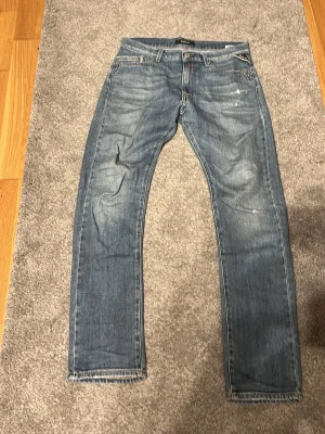 Blå Replay jeans med slitningar - Säljer ett par blå jeans från Replay med snygga slitningar och tvättade detaljer. Klassisk femficksmodell med raka ben och normal midja. Jeansen har Replay-logga på fickan och är gjorda i mjuk denim för skön passform.