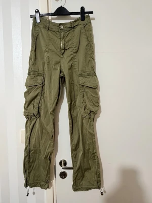 Olivgröna cargobyxor med dragsko från Guess - Snygga olivgröna cargobyxor med flera stora fickor på sidorna och dragsko vid benslut. Byxorna har avslappnad passform och klassisk cargostil, perfekta för en streetwear-look. Materialet känns lätt och bekvämt, perfekt för vardagsoutfits.