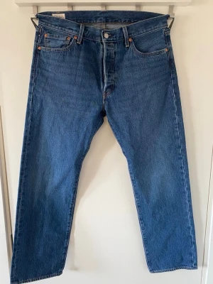 Levi's 501 jeans - Klassiska blå Levi's 501 jeans med rak passform och knappgylf. Tillverkade i slitstark bomullsdenim med fem fickor och ikonisk röd Levi's-tag på bakfickan. Perfekta för en avslappnad och tidlös stil.