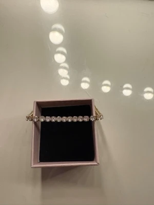 Guldigt tennisarmband med stenar - Snyggt tennisarmband i guldton med gnistrande klara stenar i rad. Armbandet har en klassisk och stilren design som passar perfekt till din outfit. Levereras i en söt rosa ask.