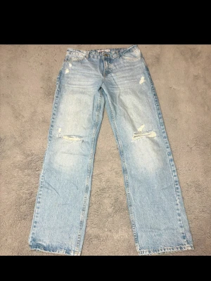 Ljusblå raka jeans med slitningar - Säljer ett par ljusblå jeans med raka ben och trendiga slitningar på låren och knäna. Jeansen har klassisk femficksdesign och knappgylf. Perfekta för en avslappnad streetstyle-look.