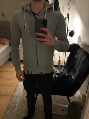 Ljusgrå zip hoodie från Ralph Lauren - Säljer en ljusgrå hoodie från Ralph Lauren med dragkedja framtill och vit dragsko i huvan. Klassisk passform med broderad logga på bröstet. Perfekt för en avslappnad och stilren look. Mjuk bomullsblandning och ribbade muddar vid ärmar och midja.