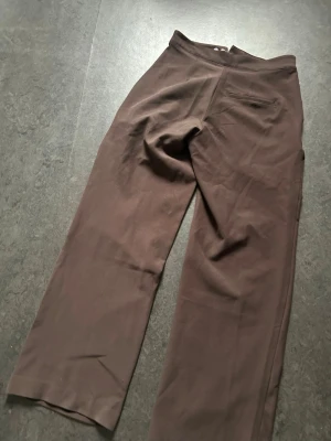 Bruna kostymbyxor med raka ben - Säljer ett par bruna kostymbyxor med raka ben och hög midja. Byxorna har diskreta fickor fram och en bakficka. Klassisk design utan synligt märke, perfekta för en clean och stilren look. Materialet ser ut att vara en mjuk och lätt tygblandning.