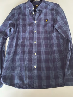 Blårutig skjorta från Lyle & Scott - Snygg blårutig skjorta från Lyle & Scott med klassisk krage och vita knappar framtill. Skjortan har lång ärm och det ikoniska gula logomärket broderat på bröstet. Perfekt för dig som gillar stilrena och tidlösa plagg.