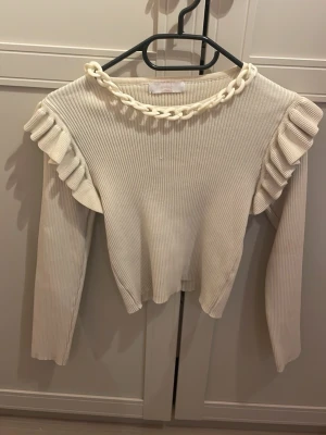Vit ribbad tröja med volang och kedja - Supersnygg vit ribbad tröja med långa ärmar och volangdetaljer längs axlarna. Tröjan har en chunky kedja runt halsringningen som ger en cool twist. Figurnära passform och mjukt material som känns skönt mot huden.