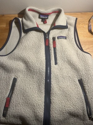 Beige fleeceväst från Patagonia - Säljer en beige fleeceväst från Patagonia med marinblå detaljer och dragkedjor. Västen har hög krage, tre fickor med dragkedja och rödbruna dragflikar. Perfekt för lager-på-lager och outdoor-vibe. Pris kan diskuteras 