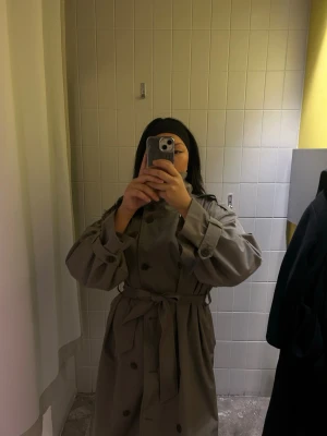 Green oversized trenchcoat - Stilren beige trenchcoat med dubbelknäppning, bred krage och knytskärp i midjan. Jackan har axelklaffar och stora knappar, samt vida ärmar med justerbara band vid handlederna. Perfekt för en trendig och avslappnad look.