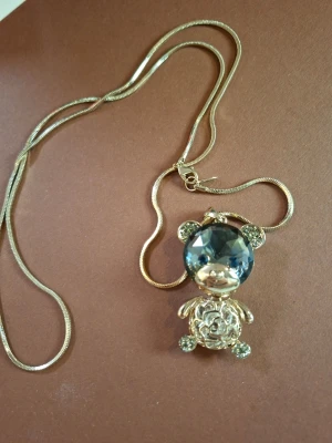 långt halsband - Unikt halsband med en guldig kedja och en stor björnberlock. Björnen har ett stort, blått glittrande huvud i kristall-look och detaljerade tassar och öron. Perfekt för dig som gillar söta och iögonfallande accessoarer.