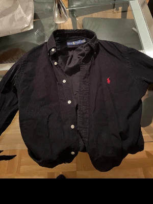 Svart manchesterskjorta från Polo Ralph Lauren - Klassisk svart manchesterskjorta från Polo Ralph Lauren med röd broderad logga på bröstet. Storlek XS