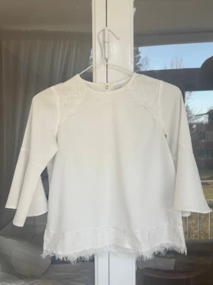 Vit blus med spetsdetaljer - Säljer en vit blus med långa ärmar och fina spetsdetaljer vid axlar och nederkant. Blusen har en rak passform och är tillverkad i ett mjukt tyg som känns skönt mot huden. Perfekt för dig som gillar en romantisk och stilren look. Varan har några få små fläckar som knappt syns.