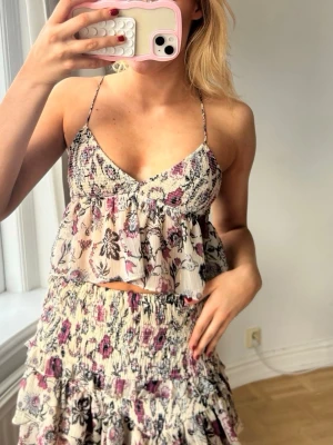 Blommig volang topp med tunna band💖 hela setet - Söt blommig topp med volang och tunna axelband. Toppen har en ljus bas med lila och svarta blommor, smockad överdel och luftig passform. Perfekt för varma dagar och ger en romantisk vibe.