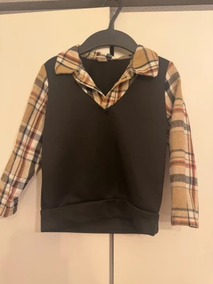 Svart tröja med rutiga ärmar och krage - Trendig svart tröja med beige, rödrutiga och vita ärmar samt matchande krage i flanell. Tröjan har en V-ringning och en avslappnad passform. Perfekt för dig som gillar lager-på-lager-looken utan att behöva flera plagg.