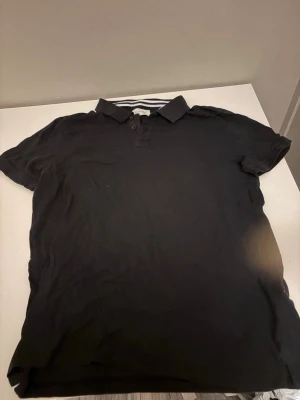 Svart pikétröja med krage - Klassisk svart pikétröja med krage och korta ärmar. Tröjan har knappar framtill och en stilren look som passar till det mesta. Perfekt för en avslappnad men ändå snygg stil.