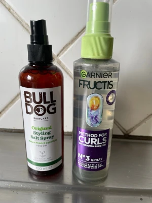 Saltvattenspray & lockspray hår - Två stylingprodukter för håret: Bulldog Original Styling Salt Spray i brun plastflaska med svart spraymunstycke och Garnier Fructis Method for Curls No3 Spray i genomskinlig plastflaska med grön spraytopp. Perfekt för att skapa textur och definiera lockar.