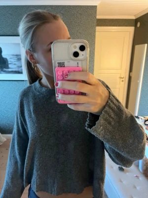 Grå croppad stickad tröja - Mysig grå stickad tröja med croppad passform och vida ärmar. Tröjan har rund halsringning och är tillverkad i ett mjukt, ull-liknande material. Perfekt för lager på lager och en avslappnad stil.