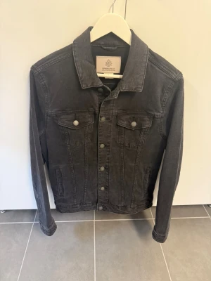 Svart jeansjacka från Springfield, stl S - Snygg svart jeansjacka från Springfield med klassisk krage, knäppning framtill och två bröstfickor med lock. Jackan har raka ärmar och en stilren look som passar perfekt till många outfits. Materialet är slitstarkt jeans och jackan har en modern passform.